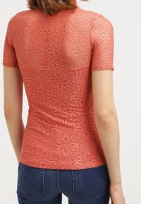 Top in pizzo corallo con maniche corte, caratterizzato da un motivo geometrico e un design testurizzato e semi-trasparente. Indossato con jeans blu scuro.