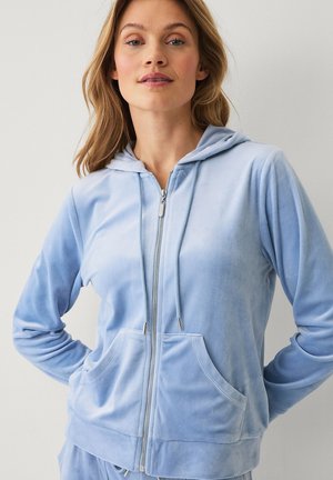 Frau mit hellbraunen Haaren, die einen hellblauen Kapuzenpullover mit Reißverschluss und Fronttaschen trägt, steht vor einem schlichten Hintergrund.