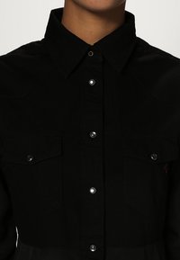 Camicia nera button-up realizzata in tessuto morbido, con colletto, bottoni a pressione e due tasche sul petto con dettagli metallici e cuciture decorative.
