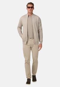 Beżowy kardigan w prążki z zamkiem z przodu, jasnobeżowa koszulka z okrągłym dekoltem oraz jasnobeżowe spodnie. Całość dopełniają brązowe trampki.