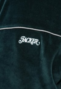 Tmavozelená sametová bunda s bielym lemovaním a vyšitým textom "JACKER" písmom v kurzíve na hornom chrbte. Jemný povrch.