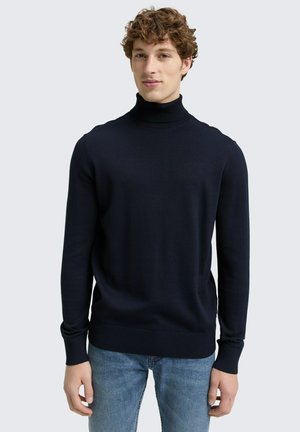Marineblauwe coltrui met ribbelkraag en -manchetten, gemaakt van gestructureerde gebreide stof, gecombineerd met lichtblauwe denim jeans.