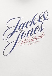 Bijela majica s velikim grafičkim natpisom u mornarsko plavoj i crvenoj boji koji glasi "Jack & Jones Worldwide Originals" u kurzivnom fontu.