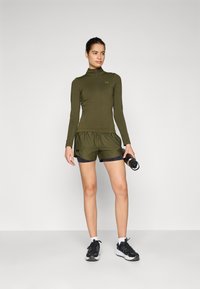 Olivengrønn idrettstopp med halv glidelås og matchende shorts. Shortsene har et lagdelt design, kombinert med svarte joggesko og en vannflaske.