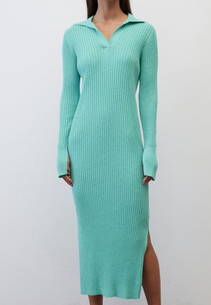 Femme portant une robe ajustée en maille côtelée bleu sarcelle à manches longues, avec un col et une fente latérale, se tenant devant un fond uni.