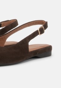 Καφέ σουέντ φλατ σανδάλι με λεπτό λουράκι στον αστράγαλο και χρυσή αγκράφα σε λευκό φόντο.