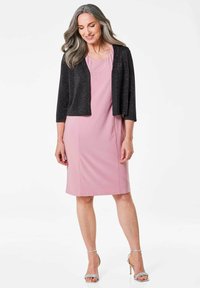 Schwarze, glitzernde, kürzere Jacke über einem hellrosa Kleid mit rundem Ausschnitt und dezenten Verzierungen am Oberteil. Silbernene Sandalen mit Knöchelriemen.