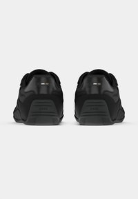 Para czarnych tenisówek widzianych z tyłu, z teksturowanymi podeszwami i subtelnymi detalami logo na zakładkach pięt i środkowej części podeszwy.