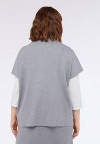 Szary dzianinowy top z krótkimi rękawami, luźną sylwetką i miękką fakturą. Noszony na białej koszuli z długim rękawem. Gładki materiał, bez wzorów.