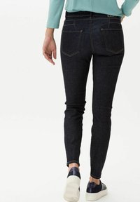 Jeans de mezclilla oscuros y ajustados de color azul con una cintura de altura media, que presentan bolsillos traseros y costuras doradas en contraste, combinados con zapatillas blancas y marino.