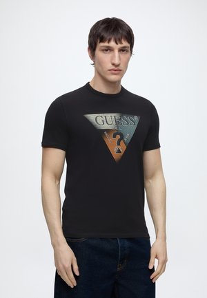 Νέος άνδρας που φοράει μαύρο μπλουζάκι Guess με μεγάλο πολύχρωμο τρίγωνο λογότυπο και σκούρο μπλε τζιν, σε απλό φόντο.