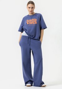 Blauwe oversized t-shirt met oranje grafische tekst, gecombineerd met bijpassende losse sweatrokken. Het materiaal lijkt zacht en heeft een relaxte pasvorm.