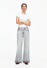 Jeans grigi ampi con una leggera trama e un lavaggio chiaro, abbinati a una T-shirt grigia aderente. Il modello posiziona le braccia incrociate.