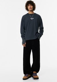 Felpa a maniche lunghe grigia scura con polsini a coste, pantaloni in velluto a coste neri e scarpe marroni con texture e lacci blu. Collana d'argento visibile.