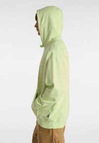 Un sweat à capuche vert clair avec un cordon de serrage, une poche kangourou et des poignets côtelés, fabriqué dans un tissu doux, porté par une personne de profil.