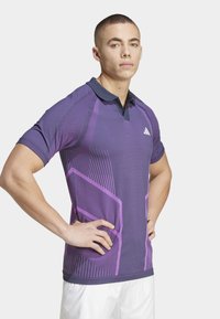 Polo deportivo morado con un cuello oscuro a contraste, que presenta rayas moradas diagonales y un tejido texturizado para transpirabilidad.