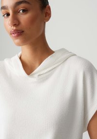 Pullover bianco con cappuccio, caratterizzato da una texture a coste, con una vestibilità morbida e maniche corte. Il tessuto sembra morbido e leggero.