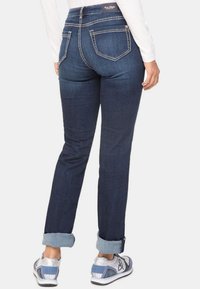 Jeans de mezclilla oscura con una silueta ajustada, que presentan una cintura de tiro medio, dobladillos enrollados y costuras en contraste en los bolsillos. Combinados con zapatillas ligeras.