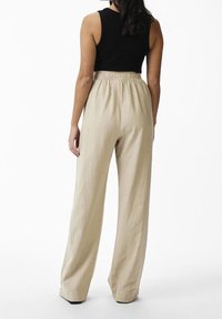 Woman wearing high-waist beige wide-leg pants and a sleeveless black top, standing facing away against a plain white background.