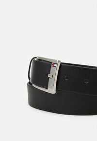 Ceinture en cuir noir avec une boucle en métal argenté ornée d'un logo coloré. La ceinture possède des trous espacés de manière égale pour un ajustement réglable.