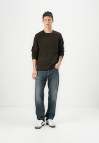 Suéter de malha texturizada verde-escuro, jeans azuis de corte largo e ténis brancos com detalhes em preto. Modelo em pé contra um fundo branco.