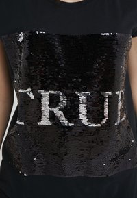 Svart t-shirt med en paljettpanel som har texten "TRUE" i vitt. Paljetterna är glänsande och tätt ordnade, vilket ger texturen.