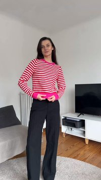 Sieviete, kura valkā rozā un baltu svītrainu džemperi un melnas bikses, stāv dzīvojamā istabā ar dīvānu un TV plauktu.