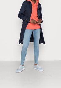 Persona con un hoodie corallo sotto un trench coat blu navy, abbinato a jeans azzurri e sneakers, in piedi su uno sfondo neutro.
