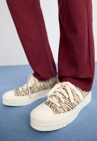 Beżowe sneakersy w czarno-białym wzorze zebry, z białą gumową osłoną na palce oraz grubą podeszwą, zestawione z burgundowymi spodniami na niebieskiej powierzchni.