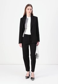 Čierny sakový blejzer a zodpovedajúce slim-fit nohavice v kombinácii s bielym topom. Ďalšie šedé akcenty na blejzeri a metalická taška.
