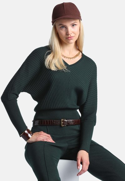 Femme blonde portant un pull côtelé vert foncé, un pantalon assorti, une casquette et une ceinture marron, un collier en or et un bracelet marron, assise avec la main sur la hanche.