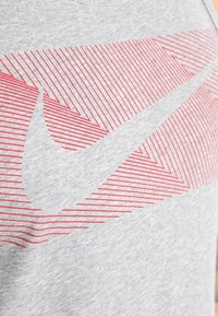 Graues Baumwoll-T-Shirt mit einem roten Nike-Logo, das diagonale Linien auf der Vorderseite bildet. Rundhalsausschnitt, kurze Ärmel.