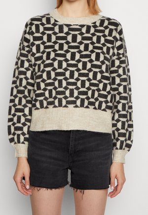 Femme portant un pull à motif géométrique noir et beige avec ourlet et poignets côtelés, associé à un short en jean noir aux bords effilochés.