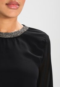 Czarna bluzka z okrągłym dekoltem, zdobiona koralikami. Gładki materiał z długimi, lekko przezroczystymi rękawami oraz luźnym krojem.
