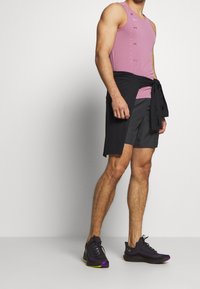 Vaaleanpunainen urheilullinen tank top, jossa on tekstuuripinta, yhdistettynä tummanharmaisiin shortseihin ja mustiin juoksukenkiin, joissa on violetteja ja vihreitä korostuksia.