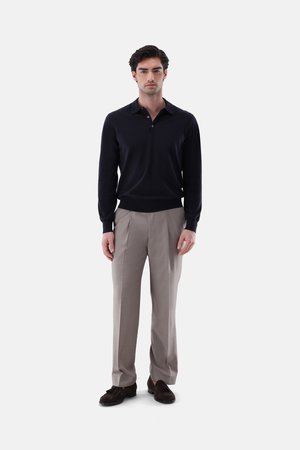 Mann steht, trägt ein schwarzes langärmeliges Poloshirt, hellbeige Hose und dunkelbraune Schuhe vor einem schlichten weißen Hintergrund.