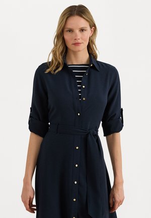 Marineblauwe jurk met opgerolde mouwen, gouden knoopdetails en een strik taille. Onder de jurk is een zwart-wit gestreepte top zichtbaar.