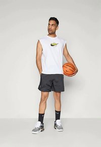 Camiseta branca sem mangas com um logótipo amarelo, calções pretos, meias pretas e ténis cinzas. Segurando uma bola de basquete laranja. De pé sobre um fundo simples.