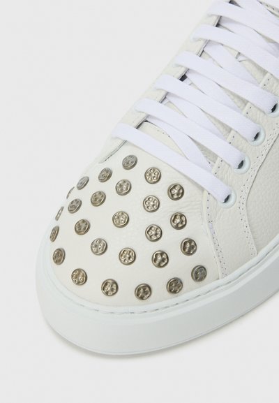 White leather sneaker with a textured upper, featuring circular metallic studs and white laces. Flat rubber sole with subtle stitching detail.