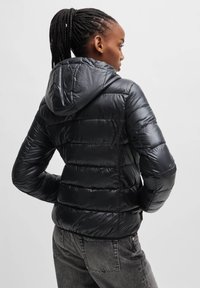 Schwarze Steppjacke mit Kapuze, mit gesteppte Abschnitten und glänzender Oberfläche. Modell ist von hinten zu sehen, kombiniert mit dunklen Jeans.