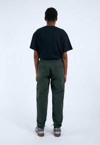 Svart bomull t-shirt och gröna byxor med elastisk midja och bakfickor. Grå och beige skor med en strukturerad sula. Bakifrån mot en ljus bakgrund.