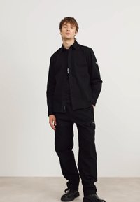 Conjunto de camiseta negra y pantalones cargo de corte relajado. Cuenta con botones, un parche con logo y tejido texturizado. Combinado con zapatos negros.