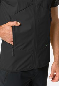 Gilet nero isolato con tasche laterali con zip e una texture liscia. Presenta cuciture angolari e una leggera lucentezza. Accent verde sulla tasca.