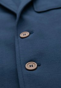 Närbild av två bruna knappar på ett mörkblått tyg, med texten "OMBRE" ingraverad på varje knapp.