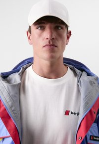 Fargerik jakke med blå, rød og grå seksjoner, med en logo. Hvit caps. Enkel hvit t-skjorte med liten "bergh" logo. Nærbilde.