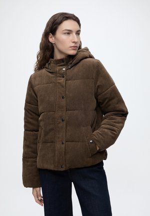 Jeune personne aux longs cheveux portant une doudoune en velours côtelé marron avec capuche et un jean foncé, debout avec une main dans la poche.