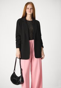 Črn blazer z dolgimi rokavi preko črne čipkaste bluze, v kombinaciji s širokimi hlačami v pastelnem roza odtenku. Drži črno lakasto torbico.