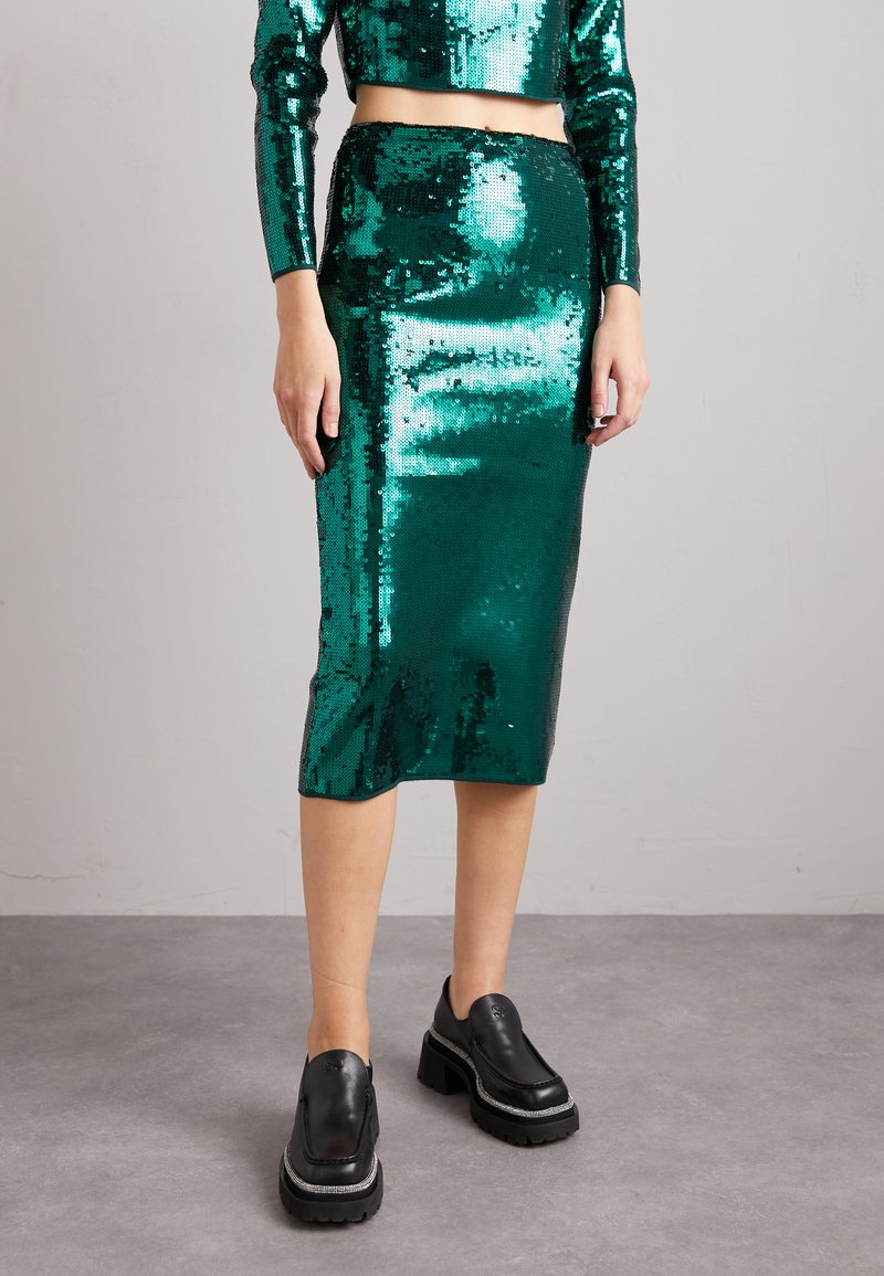 Kobieta w błyszczącej zielonej spódnicy ołówkowej pokrytej cekinami i dopasowanym krótkim topie, w połączeniu z czarnymi masywnymi loafersami na obcasie, stojąca na szarym podłożu.