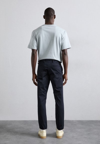 Camiseta de algodón azul claro, corte relajado, combinada con pantalones oscuros. El modelo lleva zapatos claros de tono crema con suelas de goma.