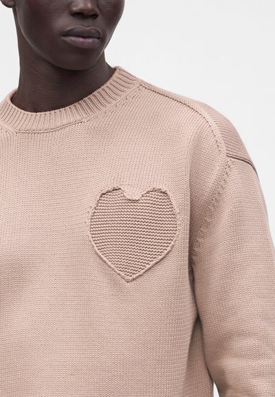 Personne à la peau foncée portant un pull en tricot rose clair avec un motif de cœur en relief sur le côté gauche de la poitrine.